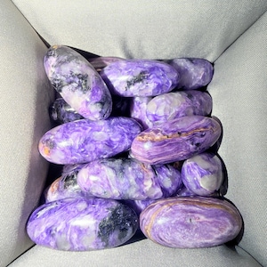 May include: A collection of polished, oval-shaped charoite stones featuring vibrant shades of purple, white, and black. Each stone displays unique swirling patterns and a smooth, glossy finish, showcasing the natural mineral texture and color variations.