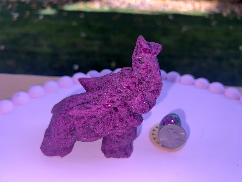 Pode incluir: Uma figura de elefante de pedra preciosa roxa em um suporte de bolo branco com uma pedra preciosa roxa menor ao lado.
