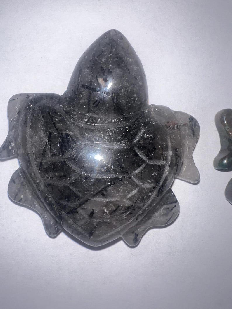May include: A carved, dark gray crystal turtle figurine with a textured surface. The turtle has a pointed head and flipper-like appendages. The crystal has black inclusions and is set against a white background.