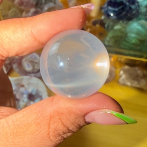 May include: A clear, iridescent crystal ball, held between fingers. The sphere has a soft, glowing interior and a smooth, polished surface. The background features other crystals and gemstones.