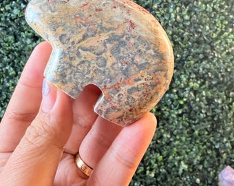 May include: A multi-colored stone carving with a rounded top and a curved cutout. The stone features a mix of beige, gray, and red hues. The carving is held in a hand against a backdrop of green foliage.