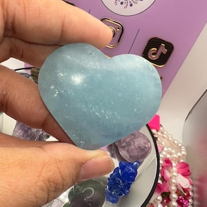 May include: A light blue heart-shaped crystal, also known as a heart chakra stone, is held in a hand. The crystal is smooth and polished, with a slight shimmer.
