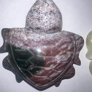 May include: A carved stone turtle figurine. The turtle's shell is a mix of red, green, and white, with a textured pattern. The head is a speckled white and gray. The figurine is a decorative item.