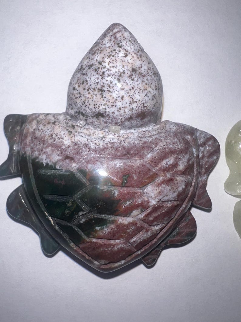 May include: A carved stone turtle figurine. The turtle's shell is a mix of red, green, and white, with a textured pattern. The head is a speckled white and gray. The figurine is a decorative item.