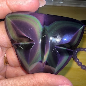 May include: A polished, iridescent obsidian butterfly carving. The stone features a blend of purple, green, and black hues. The butterfly shape is symmetrical, with detailed wing and body lines. A purple beaded bracelet is visible in the background.