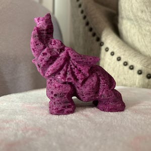 Pode incluir: Uma pequena figura de elefante esculpida em roxo, feita de um material texturizado. A figura est&aacute; em p&eacute; sobre uma superf&iacute;cie branca e fofa.