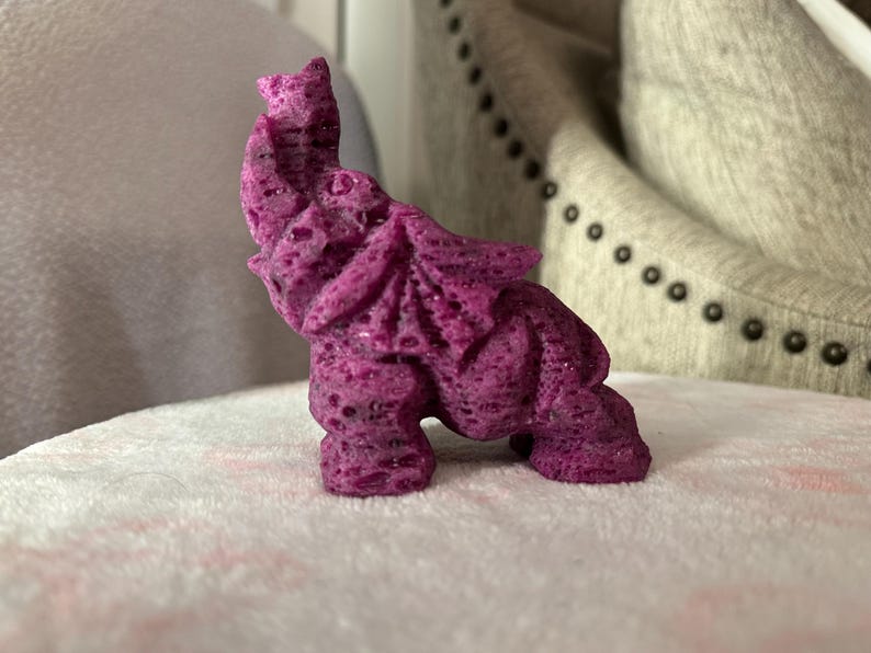 Pode incluir: Uma pequena figura de elefante esculpida em roxo, feita de um material texturizado. A figura est&aacute; em p&eacute; sobre uma superf&iacute;cie branca e fofa.