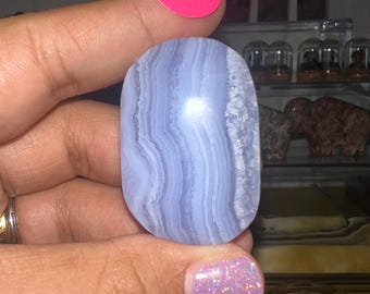 May include: A polished, oval-shaped blue lace agate gemstone. The stone features bands of light and dark blue, creating a layered effect. A pink sticker with the letter "A" is visible above the stone.
