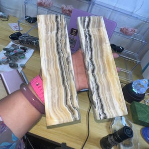 May include: Two rectangular onyx slabs with a cream and brown striped pattern. The slabs are held up, showcasing their polished surface. The background includes display cases with crystals and other stones, suggesting a collection of natural gemstones.