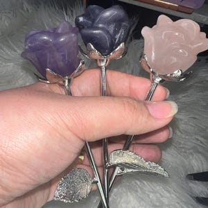 May include: Three crystal rose stems with silver metal leaves and stems. The roses are amethyst, rose quartz, and a dark purple color.