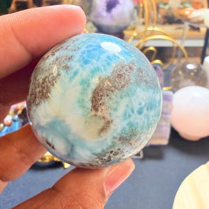 Op de afbeelding: Een gepolijste, bolvormige Larimar-edelsteen met een levendige hemelsblauwe kleur, ingewikkelde witte marmering en vlekken van aardse bruine minerale insluitingen. Het oppervlak is glad en reflecterend en toont overal natuurlijke patronen.