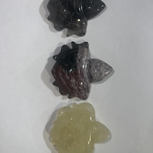 May include: Three carved stone figurines in the shape of a fish. The top figurine is dark gray, the middle one is a mix of red, gray, and white, and the bottom one is a pale yellow color. The figurines are displayed on a white background.