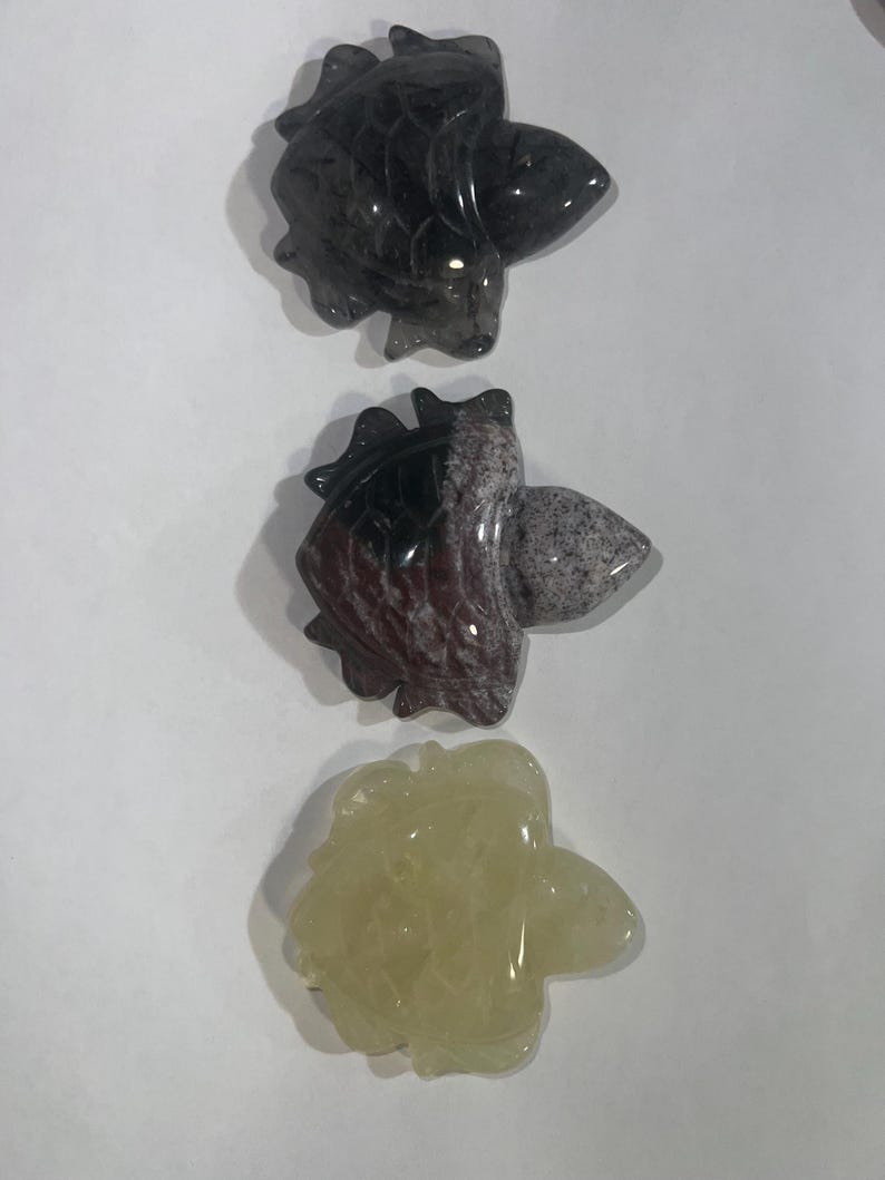 May include: Three carved stone figurines in the shape of a fish. The top figurine is dark gray, the middle one is a mix of red, gray, and white, and the bottom one is a pale yellow color. The figurines are displayed on a white background.