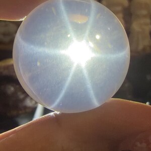 May include: A translucent, spherical gemstone with a six-rayed star pattern. The gem is held between fingers, showcasing its polished surface and the star's bright, radiating lines. The stone has a soft, milky appearance.