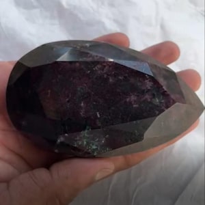 May include: A large, faceted gemstone with a pear shape. The gem has a dark, translucent exterior with a deep burgundy interior. The facets catch the light, creating a sparkling effect. The gemstone is held in a hand.
