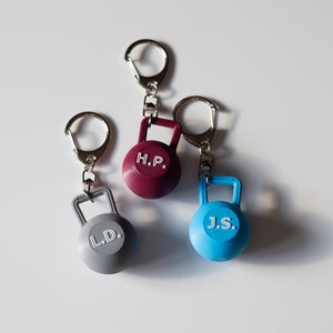 Könnte beinhalten: Drei kleine, farbenfrohe, schlüsselanhängergroße Kettlebells aus Metall mit silbernen Schlüsselringen. Die Kettlebells sind rot, grau und blau. Die rote Kettlebell trägt die Buchstaben "H.P.", die graue Kettlebell die Buchstaben "L.D." und die blaue Kettlebell die Buchstaben "J.S.".
