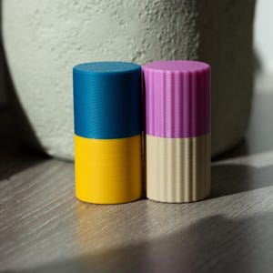 Op de afbeelding: Twee cilindrische containers met deksels. De container aan de linkerkant is blauw en geel. De container aan de rechterkant is roze en beige.