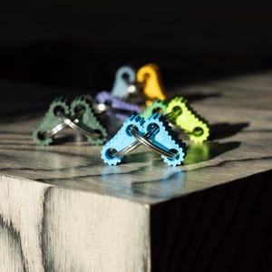Puede incluir: Una colección de llaveros coloridos en tonos azul, verde, amarillo y morado. Cada llavero presenta un diseño único en forma de engranaje con un llavero plateado. Los llaveros están dispuestos sobre una superficie de madera gris, con sombras.