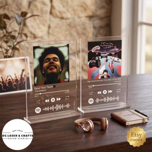 May include: Two clear acrylic music plaques featuring album art and song details. One displays "Save Your Tears" by The Weeknd, the other "High School Reunion" by SAINt JHN. A pair of rose gold headphones and a small notebook are also visible.