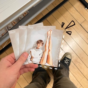 May include: Three square cards with the word "Hers" printed on them. Each card features an illustration of a person in a white sweater and a pair of legs. The cards are held in a hand.