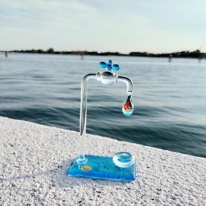 Op de afbeelding: Een heldere glazen kraansculptuur met een blauwe vlindervormige handgreep en een blauwe basis. Een druppelvormig glazen element hangt aan de kraan en bevat een kleurrijk ontwerp. De sculptuur staat tegen een vage waterachtergrond.