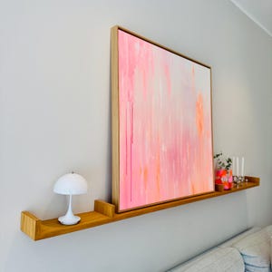 Könnte beinhalten: Ein modernes Wohnzimmer mit einem großen abstrakten Gemälde in Rosa- und Orangetönen, eingerahmt in Naturholz. Eine weiße Pilzlampe und dekorative Gegenstände befinden sich auf einem Holzregal unter dem Kunstwerk.
