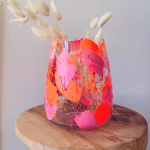 Könnte beinhalten: Eine transparente Glasvase mit einem lebendigen abstrakten Design in Rosa- und Orangetönen. Die Vase ist mit getrockneten, flauschigen, beigefarbenen Blumen gefüllt. Die Vase steht auf einer Holzoberfläche.