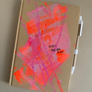 Könnte beinhalten: Ein braunes Notizbuch mit abstrakten neonrosa und orangefarbenen Pinselstrichen und dem Text "Honey, your soul is art". Ein weißer Stift ist mit einem weißen Gummiband am Notizbuch befestigt. Das Notizbuch ist geschlossen.