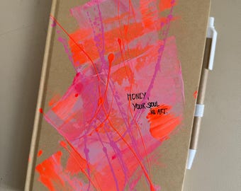 May include: A brown notebook with abstract neon pink and orange paint strokes and the text "Honey, your soul is art." A white pen is attached to the notebook with a white elastic band. The notebook is closed.