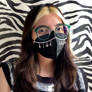 May include: A black face mask with a silver chain and star charms is worn with green-framed glasses. The background is a black and white zebra print. The person is wearing a black top with silver sequins.