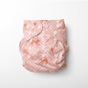 Può includere: Un pannolino di stoffa rosa con un motivo a scacchi e un design ripetitivo di cervi e aquiloni. Il pannolino ha bottoni a pressione bianchi.