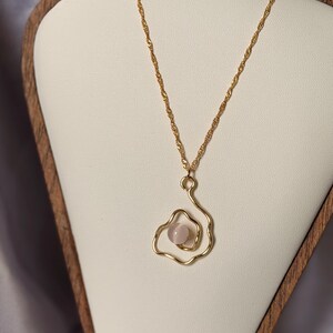 Puede incluir: Un collar de tono dorado con una cadena trenzada fina y un colgante compuesto por una cuenta esférica de color rosa pálido, lisa, encerrada en un marco de alambre dorado ondulado e irregular que forma una estructura abierta y fluida.
