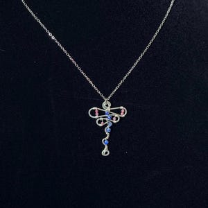 May include: A silver-toned wire-wrapped pendant shaped like a dragonfly, suspended from a thin silver chain. The pendant features small, round pink and blue beads integrated into the wire structure, creating a delicate and intricate design.