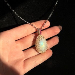 May include: A teardrop-shaped moonstone pendant encased in intricate gold wire wrapping, featuring a small pink accent bead at the top bail. The pendant hangs from a delicate gold chain, displaying an iridescent sheen on the stone surface.