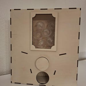 May include: A light-coloured wooden vending machine with a rectangular clear window displaying several small, round, transparent plastic capsules inside. The front panel features a circular dial and a dispensing opening at the bottom.