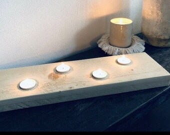 May include: A wooden candle holder with four lit tea light candles. The rectangular holder is made of light-colored wood. A lit pillar candle and a decorative vase are also visible.