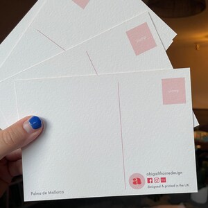 May include: A white postcard with a pink stamp area and the text "Palma de Mallorca". The postcard has a logo with the letter "a" and the text "abigailthornedesign" with social media icons for Facebook, Instagram, and Etsy. The text "designed & printed in the UK" is also included.