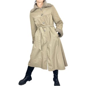 May include: A tan trench coat with a knit collar and cuffs. The coat features a belted waist, button closures, and pockets. The long coat is knee-length and has a flared skirt. The model is wearing black boots.
