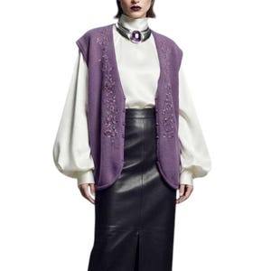 May include: A purple sleeveless knit vest with floral embroidery and button closures, worn over a white long-sleeve blouse with a high collar. A silver choker with a large purple stone is worn at the neck. A black leather midi skirt with a front slit.