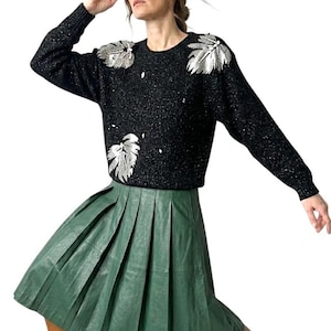 Può includere: Un maglione nero con abbellimenti argentati a forma di foglie sulle spalle e sul petto. È abbinato a una gonna verde a pieghe. L'outfit è completato da stivali marroni.