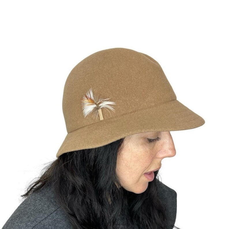 Pode incluir: Um chap&eacute;u cloche bege com uma aba pequena e um detalhe decorativo de penas. O chap&eacute;u &eacute; feito de um material macio e tem uma copa arredondada. O detalhe de penas apresenta penas marrons, brancas e bege.