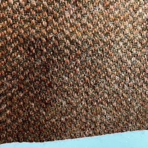 Puede incluir: Una vista de cerca de una tela tejida con un patr&oacute;n denso y texturizado. El material muestra una mezcla de fibras de color marr&oacute;n oscuro y naranja quemado, creando un tejido geom&eacute;trico repetitivo y consistente que cubre toda la superficie visible.