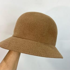 Pode incluir: Um chap&eacute;u cloche castanho com uma copa arredondada e uma aba inclinada para baixo. O chap&eacute;u &eacute; feito de um material macio semelhante a feltro. A aba tem aproximadamente 5 cm de largura.