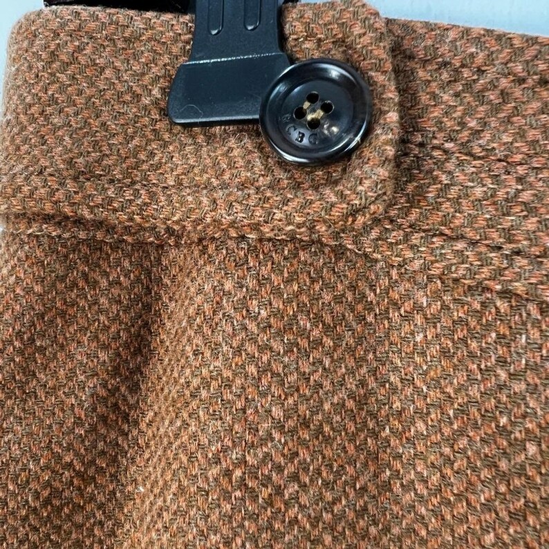 Puede incluir: Una prenda de tela texturizada marr&oacute;n que presenta una cinturilla con un solo bot&oacute;n marr&oacute;n oscuro. El bot&oacute;n est&aacute; grabado con las letras BCBG. La tela tiene un patr&oacute;n tejido visible con una mezcla de hilos de color marr&oacute;n claro y oscuro en todo el material.