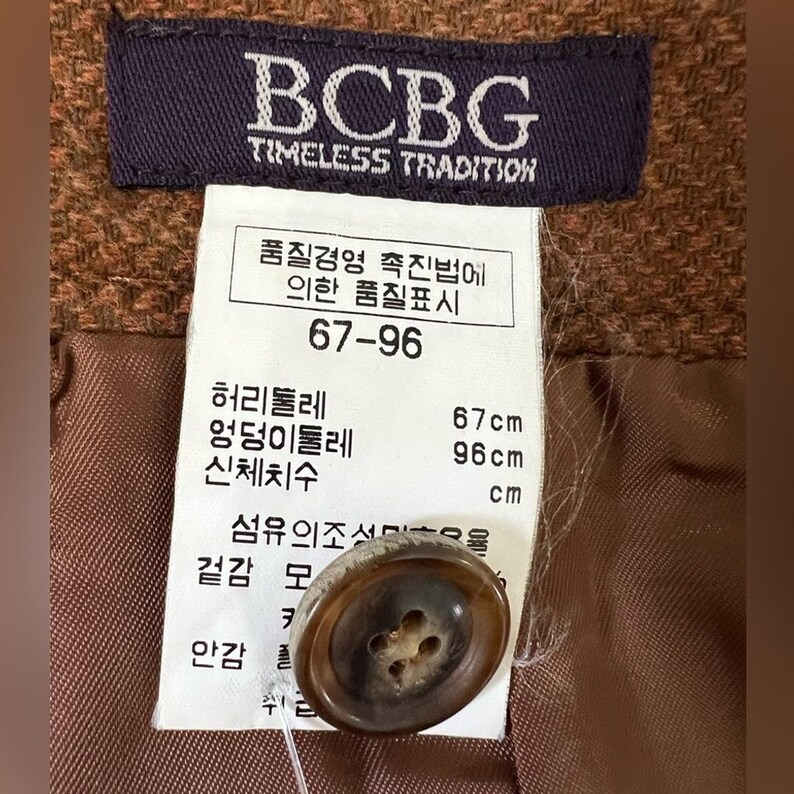 Puede incluir: Una prenda texturizada de color marr&oacute;n con una etiqueta de marca morada oscura BCBG Timeless Tradition y una etiqueta blanca con texto en coreano, incluyendo medidas de 67 cm y 96 cm, junto a un bot&oacute;n marr&oacute;n redondo de cuatro agujeros.