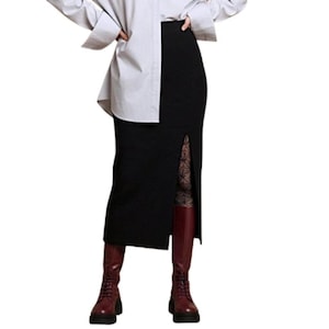 May include: A black midi skirt with a front slit, worn with a light grey button-down shirt, patterned tights, and dark red leather boots. The skirt has a textured fabric and a straight silhouette that falls below the knees.