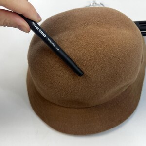 Pode incluir: Um chap&eacute;u cloche castanho com uma coroa arredondada e uma aba. Uma caneta preta com o texto "Paper Mate Write Bros 1.0" &eacute; segurada perto do chap&eacute;u. O chap&eacute;u est&aacute; sobre uma superf&iacute;cie branca.