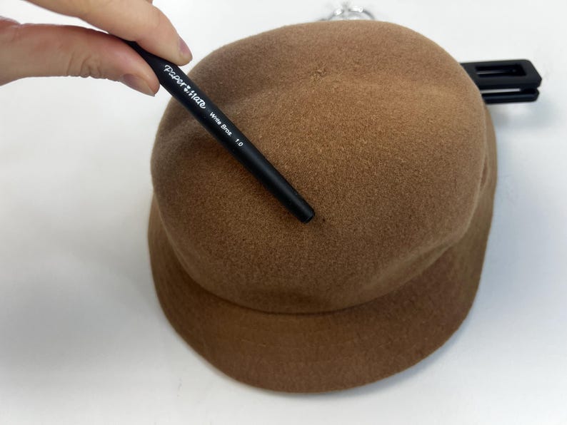 Pode incluir: Um chap&eacute;u cloche castanho com uma coroa arredondada e uma aba. Uma caneta preta com o texto "Paper Mate Write Bros 1.0" &eacute; segurada perto do chap&eacute;u. O chap&eacute;u est&aacute; sobre uma superf&iacute;cie branca.