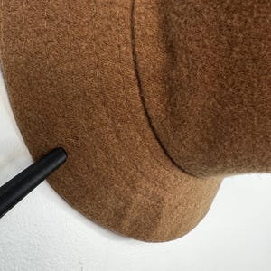 Pode incluir: Um close-up de um chap&eacute;u de l&atilde; castanho. O chap&eacute;u tem uma copa arredondada e uma aba. O chap&eacute;u &eacute; feito de um material macio e texturizado. O chap&eacute;u &eacute; de uma cor quente e neutra.