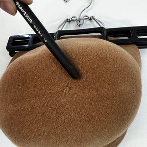 Pode incluir: Uma boina castanha pendurada num cabide preto. Uma caneta preta com o texto "Paper Mate Write Bros 1.0" aponta para a boina. A boina &eacute; feita de um material macio e felpudo.